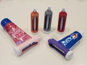 Tubes of toothpaste with a small, round device with a knob is attached to the bottom of the tube. In the background are examples of the device standalone.